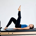 A professional female Pilates instructor in a modern, brightly lit studio. She is demonstrating a precise and controlled Pilates exercise on a reformer, showcasing perfect form and natural pose. Her movements are fluid and strong. She wears modest, professional athletic wear, fully clothed and appropriate attire. The studio is clean with natural light. The image has perfect anatomy, correct proportions, well-formed hands, and proper finger count. It is a high-quality professional photography, safe for work, appropriate content, and family-friendly.
